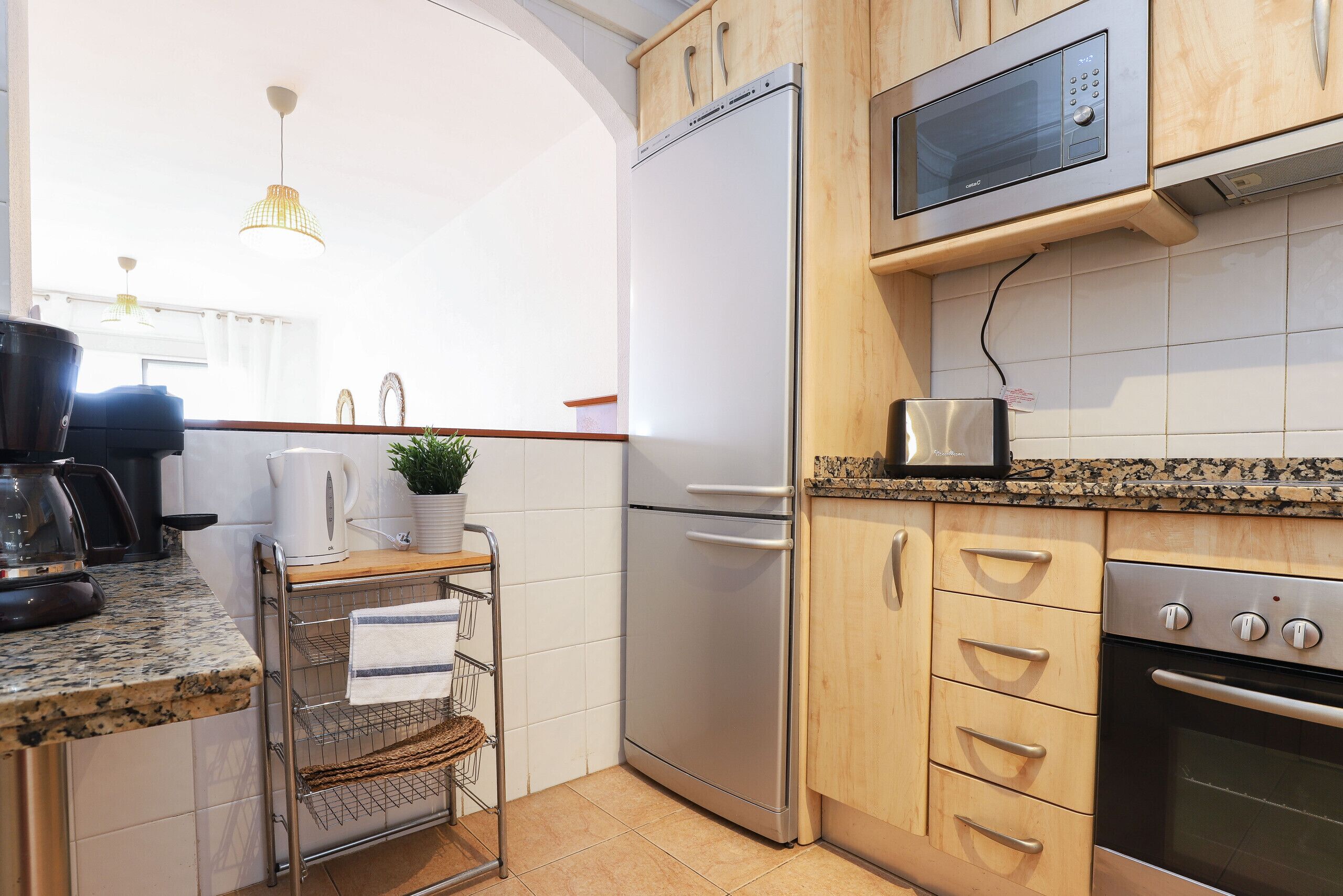 Eine kompakte Küche mit Edelstahl-Kühlschrank, Mikrowelle und Backofen. Es gibt eine kleine Arbeitsfläche mit einem Wasserkocher und einem Blumentopf.