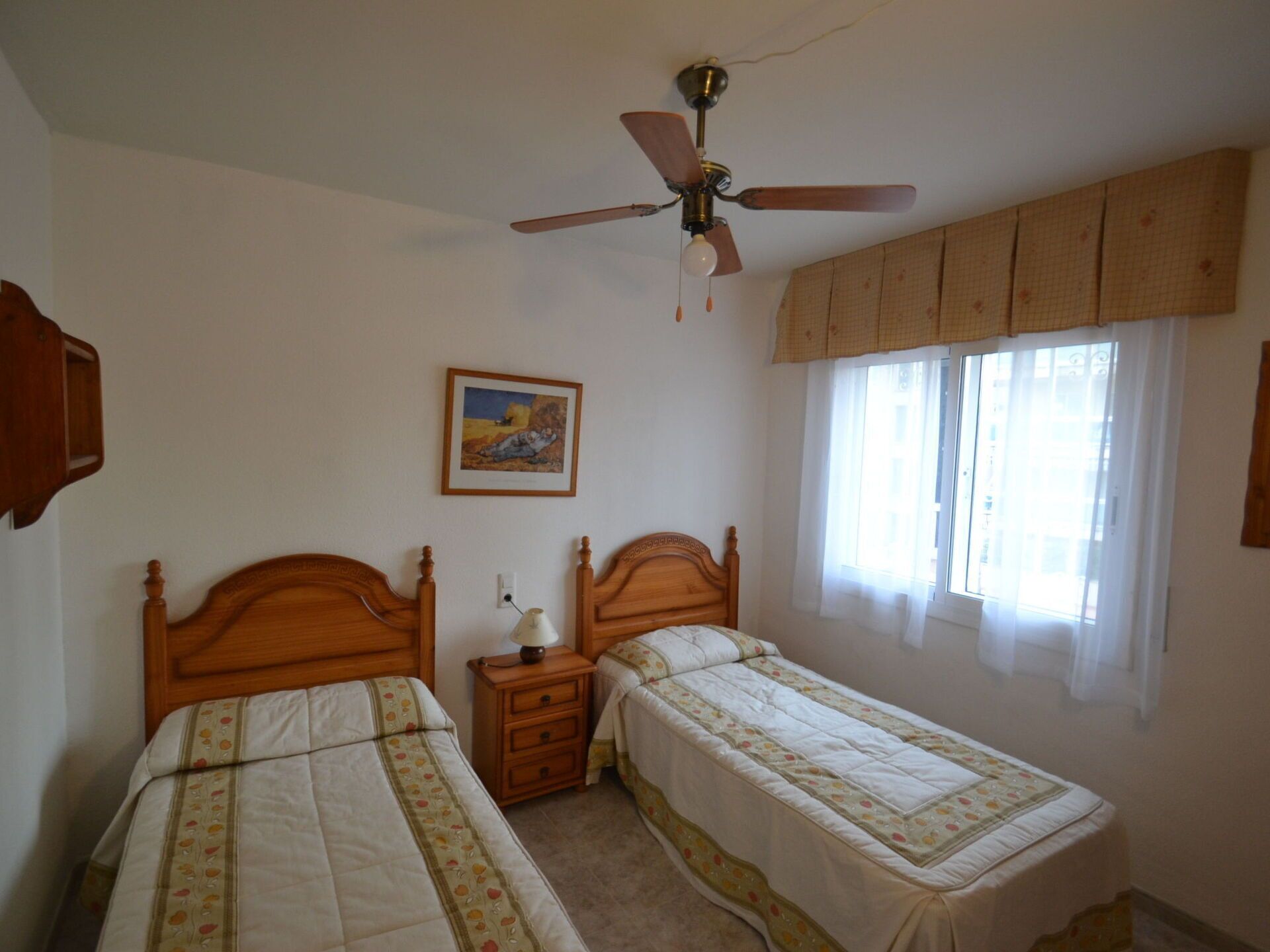 Une chambre à coucher avec deux lits en bois, un ventilateur de plafond, une table de chevet avec une lampe, une fenêtre avec des rideaux et un tableau encadré au mur.