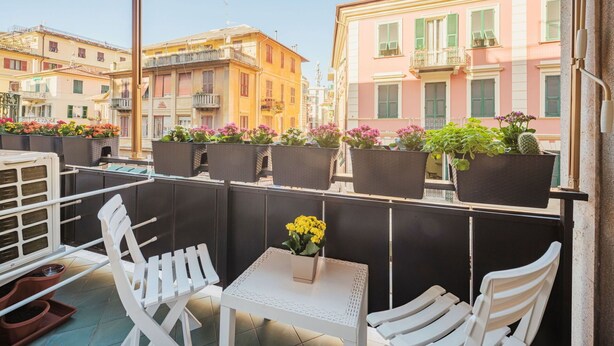A balcony with white chairs and a small table, potted plants, and a view of colorful buildings. - Cosy apartment for 2 guests with A/C, WIFI, TV and balcony (Rapallo)