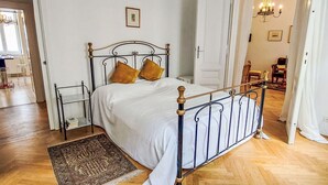 A bedroom with a metal bed frame, white bedding, two pillows, a wooden floor, a patterned rug, and a chandelier in the adjacent room. - Nice apartment for 4 people with WIFI, TV and balcony (Vienna)