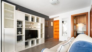 Una sala de estar moderna con un sofá, una estantería blanca y una cocina visible a través de un umbral abierto.