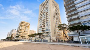 Un edificio de gran altura frente al mar con varios balcones, una playa arenosa en primer plano y un cielo azul despejado.