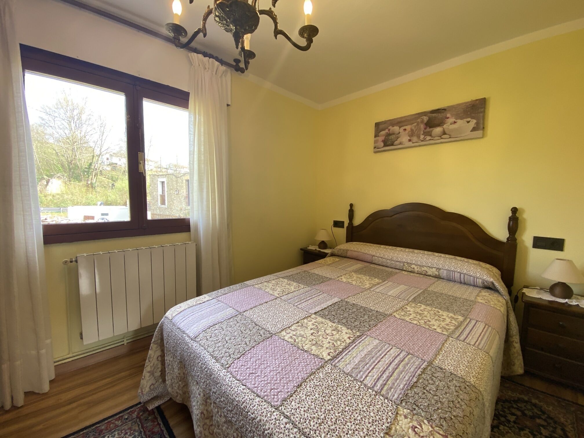 A bedroom with a large bed, a window with curtains, a radiator, a wooden headboard, a nightstand with a lamp, and a painting on the wall.