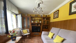 Sala de estar com sofá cinza, almofadas amarelas, armário de madeira com portas de vidro, televisão e planta em vaso sobre uma mesa.