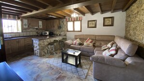 A cozy living area with a stone fireplace, a sofa, and a coffee table. There is a kitchen area with wooden cabinets and a stone island. - Casa El Molín 3 Soto de Dueñas (Soto)