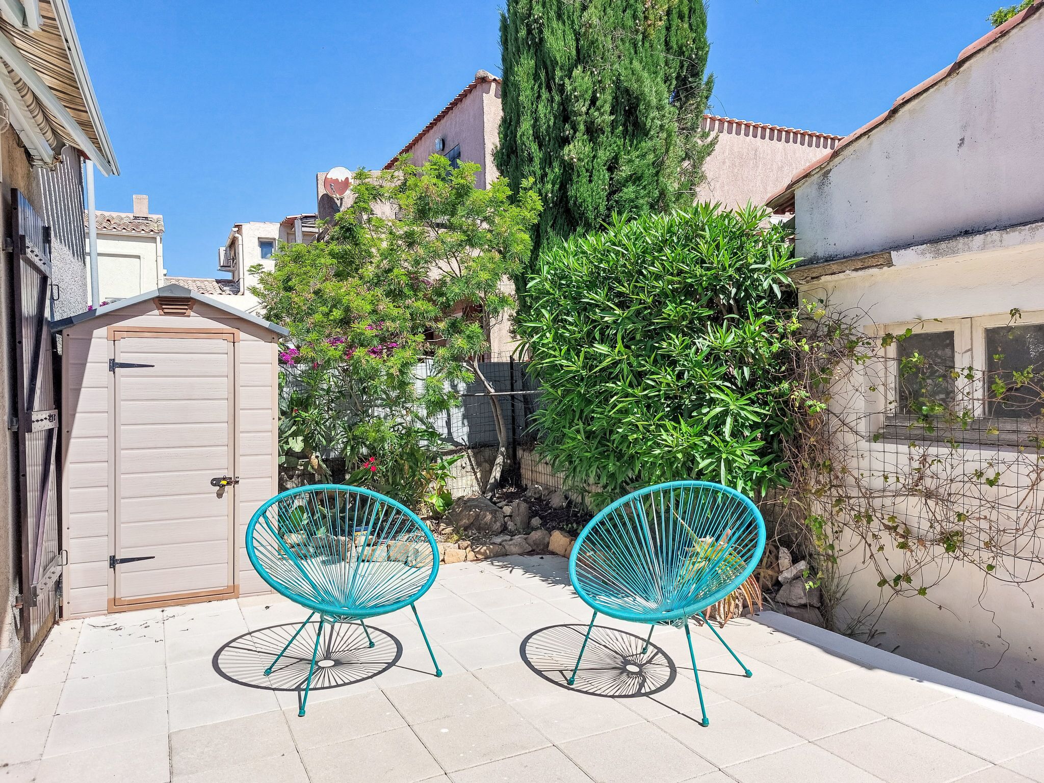 A narrow outdoor space with two turquoise chairs, a wooden gate, and a tall tree.