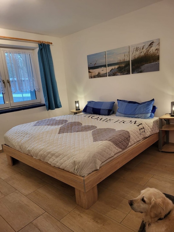 Un dormitorio con cama, una ventana con cortinas, una mesita de noche, una lámpara y un perro.