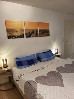 Een slaapkamer met een bed, twee aan de muur hangende afbeeldingen, een nachtkastje met een lamp en een deur.