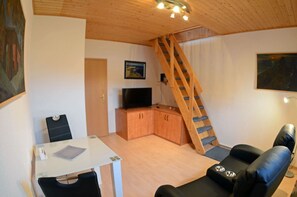Kompakti olohuone, jossa on ruokapöytä, sohva, televisio ja puinen portaikko.