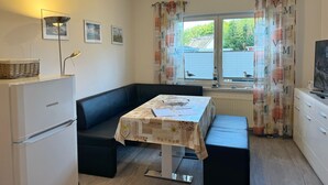 Eine kleine Küche mit Kühlschrank, einem Esstisch mit floralem Tischläufer und einem Fenster mit Vorhängen.