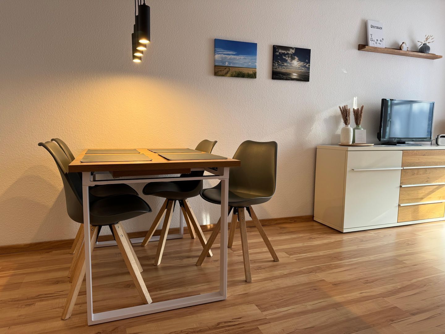 A modern dining area with a wooden table, four chairs, a hanging light fixture, and a television on a cabinet.