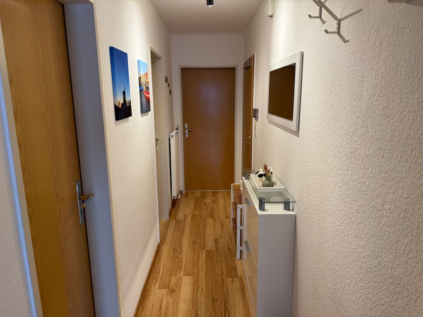 Um corredor estreito com chão de madeira, paredes brancas e uma porta de madeira no final. Há duas molduras com quadros na parede e uma pequena estante com um vaso e objetos decorativos.