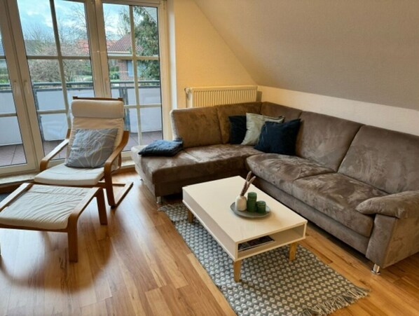 Een woonkamer met een bruine hoekbank, een houten salontafel, een witte fauteuil en een kamerkleed met dessin.