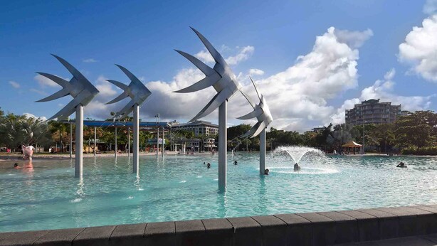 Ein Schwimmbecken mit großen Windmühlenschulpturen, umgeben von einem gepflasterten Weg und Gebäuden im Hintergrund.