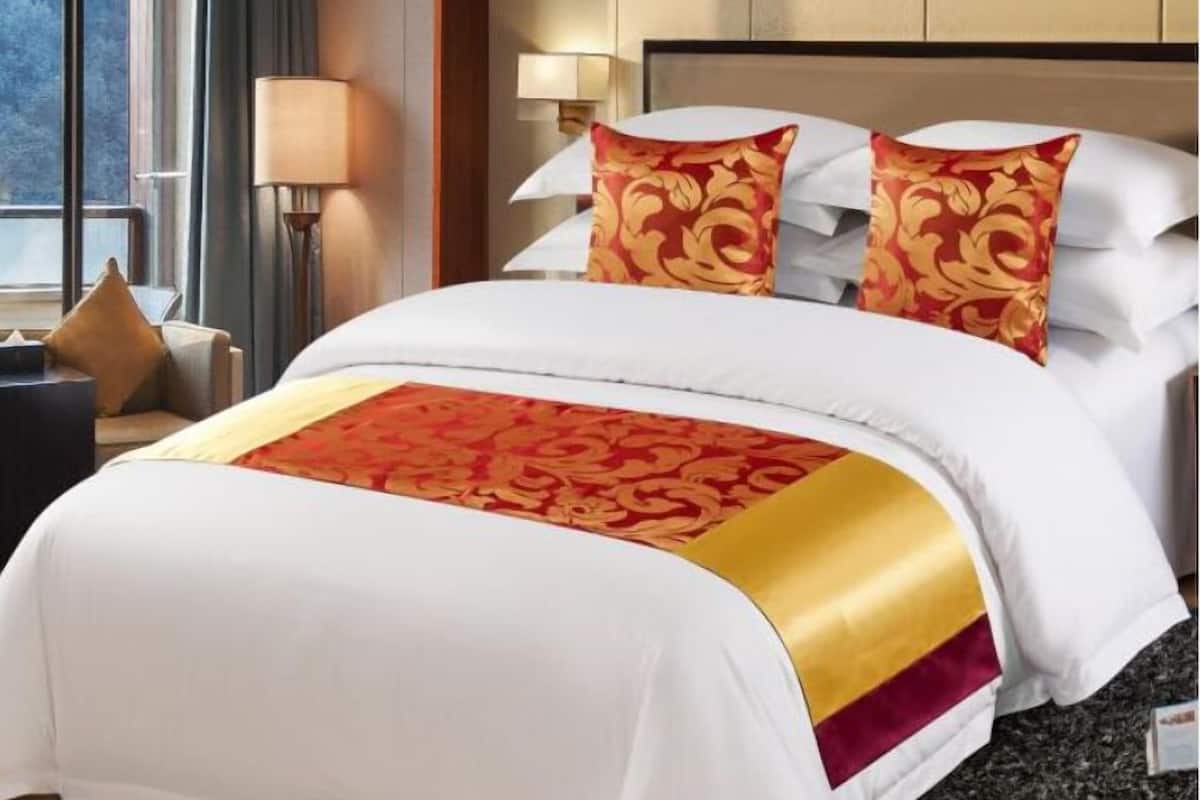 Una habitación de hotel con una cama grande, ropa de cama blanca y una manta con estampado rojo y dorado.