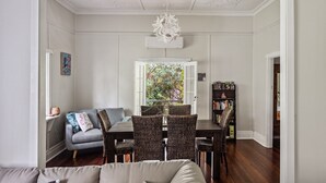 Een eetgedeelte met een tafel en stoelen, een bank, een boekenkast en een raam met uitzicht op groen.