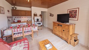 A cozy living room with a wooden ceiling, a dining area with a table set for four, a television on a wooden cabinet, and a kitchen in the background. - Wonderful apt 4/6 ppl, mountain view (Les Deux Alpes)