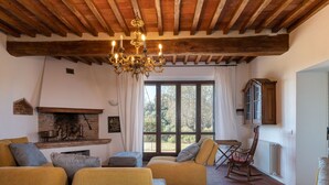 Ein gemütliches Wohnzimmer mit Kamin, Holzbalken und einem großen Fenster mit Blick auf Bäume.