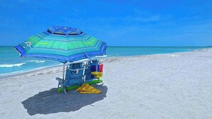 Ein Strandbienenkorb mit blau-grün gestreiftem Schirm, ein Liegestuhl und eine Kühlbox im Sand.