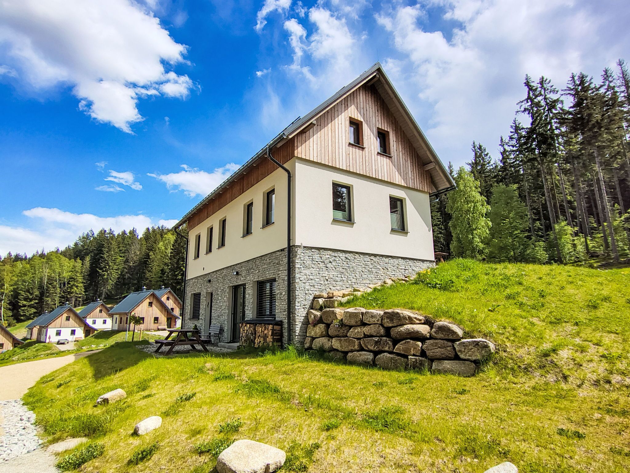 Une maison en bois à deux étages, avec des fondations en pierre, entourée d’une colline herbeuse et d’une forêt.