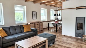Una sala moderna con un sofá, un reposapiés y una mesa de centro de madera. Hay una zona de cocina con una mesa de comedor y sillas, y una chimenea.