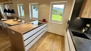 Una cocina moderna con una isla de madera, muebles blancos y encimeras negras. Tres ventanas ofrecen vistas al exterior.