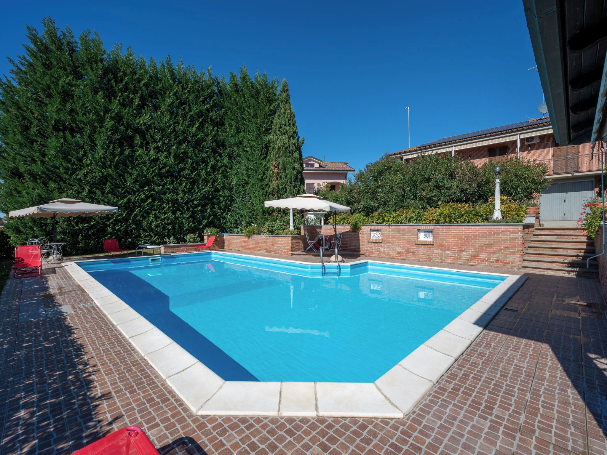 A rectangular swimming pool with a tiled edge, surrounded by a brick patio. There are two umbrellas providing shade, and a brick wall with a small window.