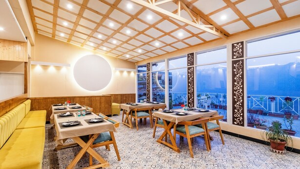 Un restaurante con mesas y sillas de madera, un sofá amarillo y ventanas amplias con vistas.