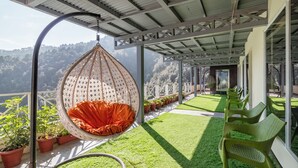 Una silla colgante con cojín rojo, sillas verdes, plantas en macetas y vista a un paisaje montañoso.