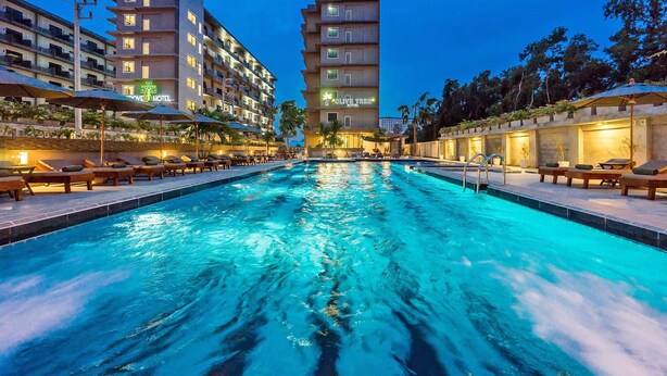 A hotel with a swimming pool, sun loungers, and a well-lit building facade. - Olive Tree Hotel (Sattahip)