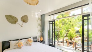 A bedroom with a large bed, two wall-mounted leaves, a nightstand, and a view of an outdoor patio with plants and furniture. - Luna Verde Hotel Hoi An (Da Nang)