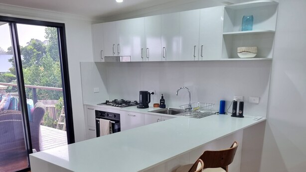 A modern kitchen with white cabinets, a central island with bar stools, a built-in oven, and a large window overlooking a deck. - Yungaburra Whitehouse 
Walking distance to village (100 metres)
2 ensuites (Yungaburra)
