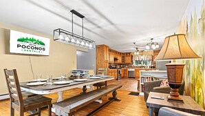 Una cocina amplia con armarios de madera, un comedor con una mesa grande y bancos, y un rincón de estar acogedor con una lámpara.