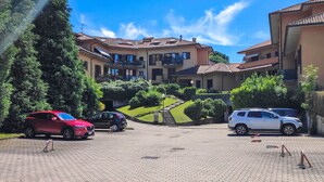 A courtyard with a paved area, a multi-story building with balconies, and parked cars. - Haridian by Interhome (Porto Valtravaglia)