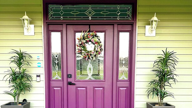 A purple front door with a decorative wreath, flanked by two potted plants, and a welcome mat on the porch.