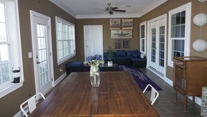 A dining area with a wooden table, white chairs, a vase with flowers, a ceiling fan, and a sofa with blue cushions.