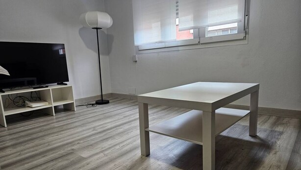 Una habitación con una mesa blanca, un televisor sobre un mueble, una lámpara de pie y una ventana con persianas.