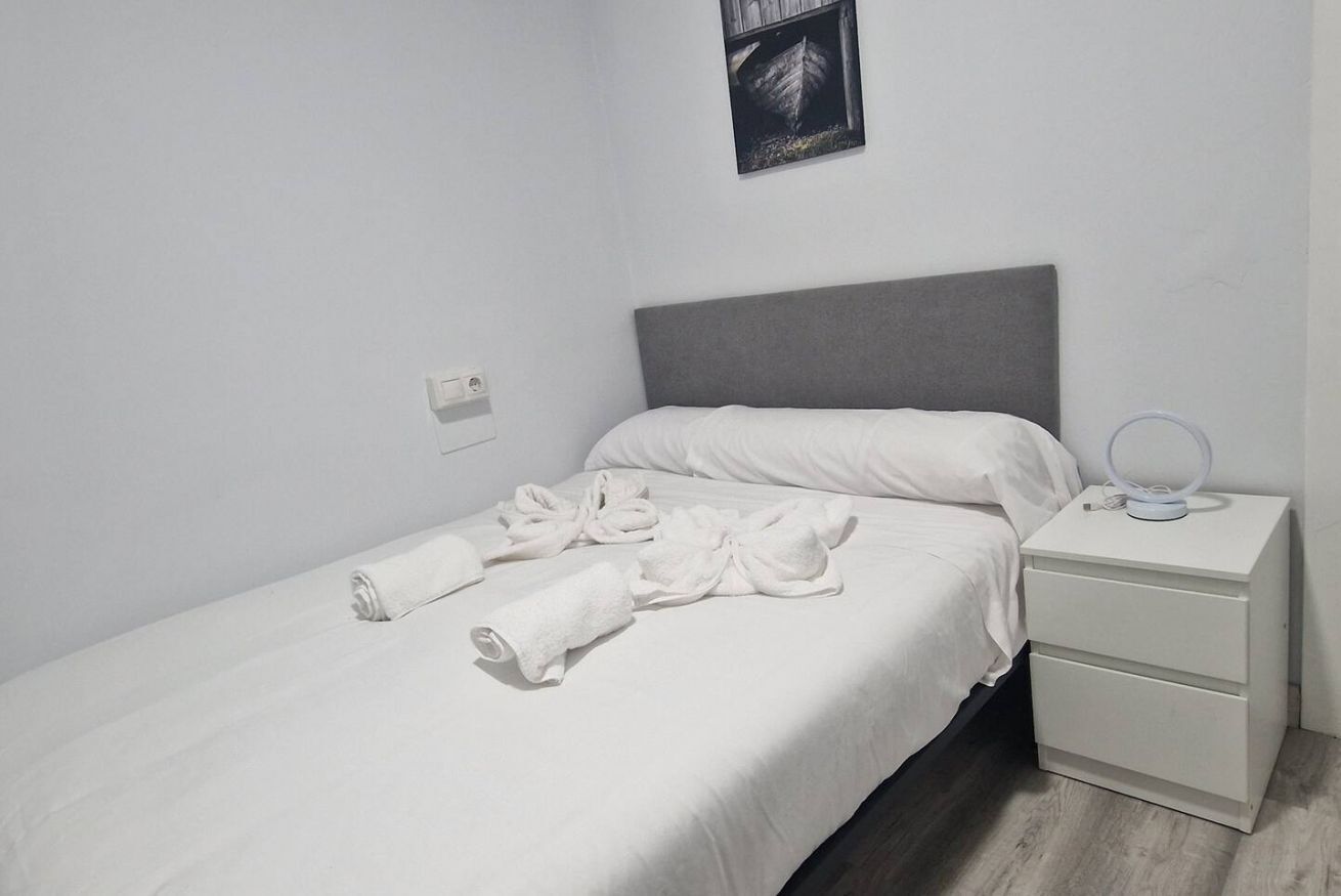 Een eenpersoonsbed met witte lakens, een grijs hoofdbord, een nachtkastje met een lamp, en een aan de muur hangend schilderij.
