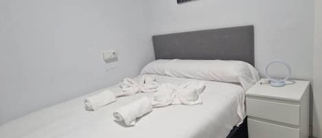 A single bed with white linens, a grey headboard, a bedside table with a lamp, and a wall-mounted picture.