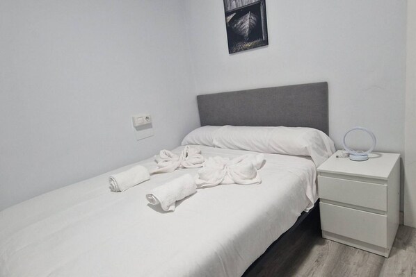 A single bed with white linens, a grey headboard, a bedside table with a lamp, and a wall-mounted picture.