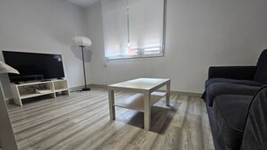 Una sala de estar con un televisor de pantalla plana, una lámpara de pie, una mesa blanca y un sofá oscuro.