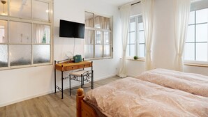 A bedroom with two beds, a desk with a chair, a television, and large windows with curtains.