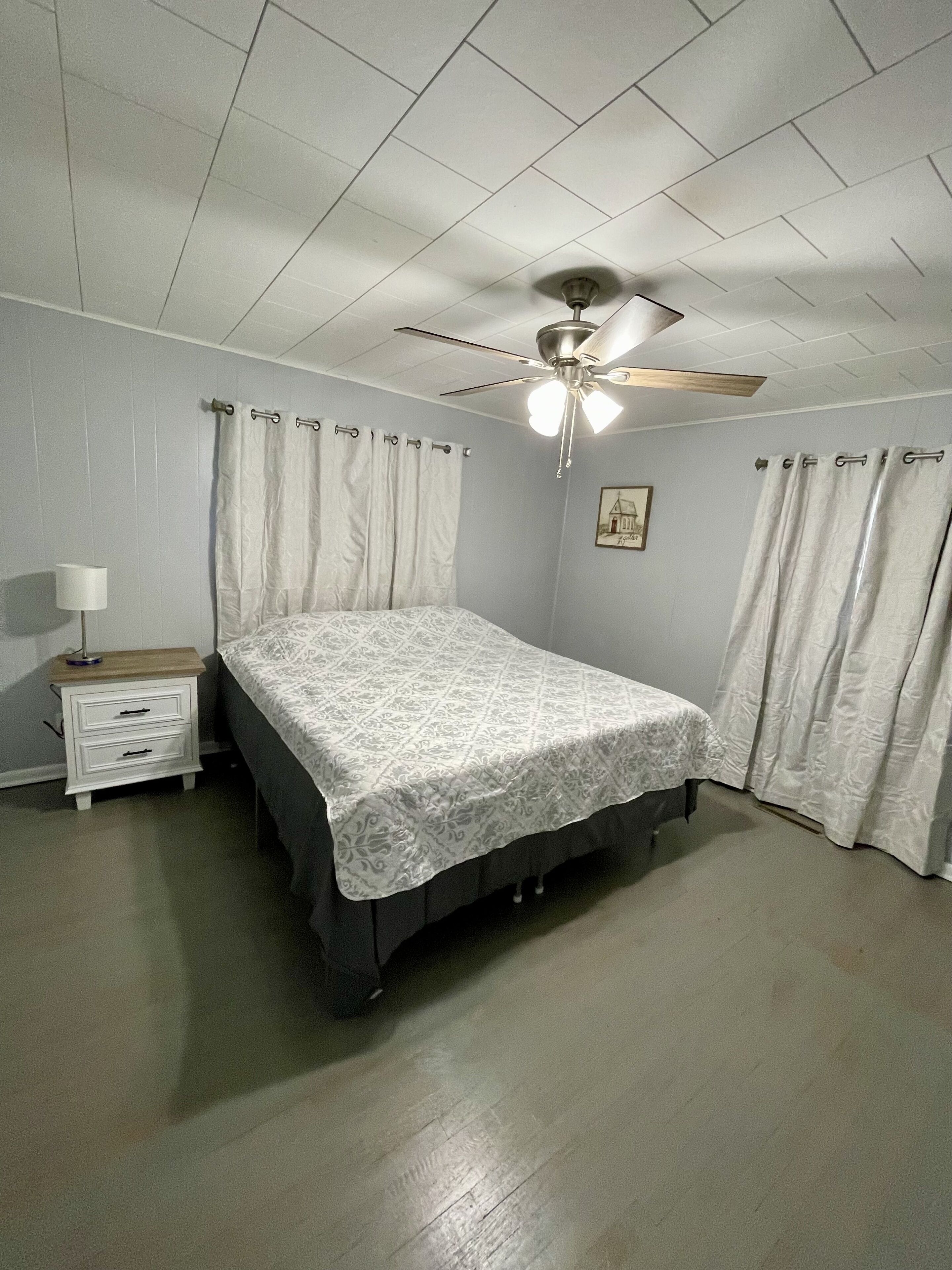 A bedroom with a bed, a nightstand, a ceiling fan, and light-colored curtains.