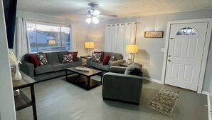A living room with a sectional sofa, a loveseat, a coffee table, and two lamps. There is a door with a 'Blessed' sign above it and a window with curtains.