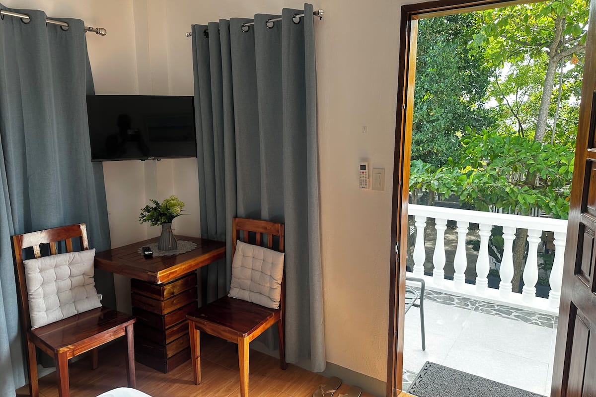 Una habitación con un escritorio de madera, dos sillas, una mesita con un florero y vista a la vegetación por una puerta abierta.