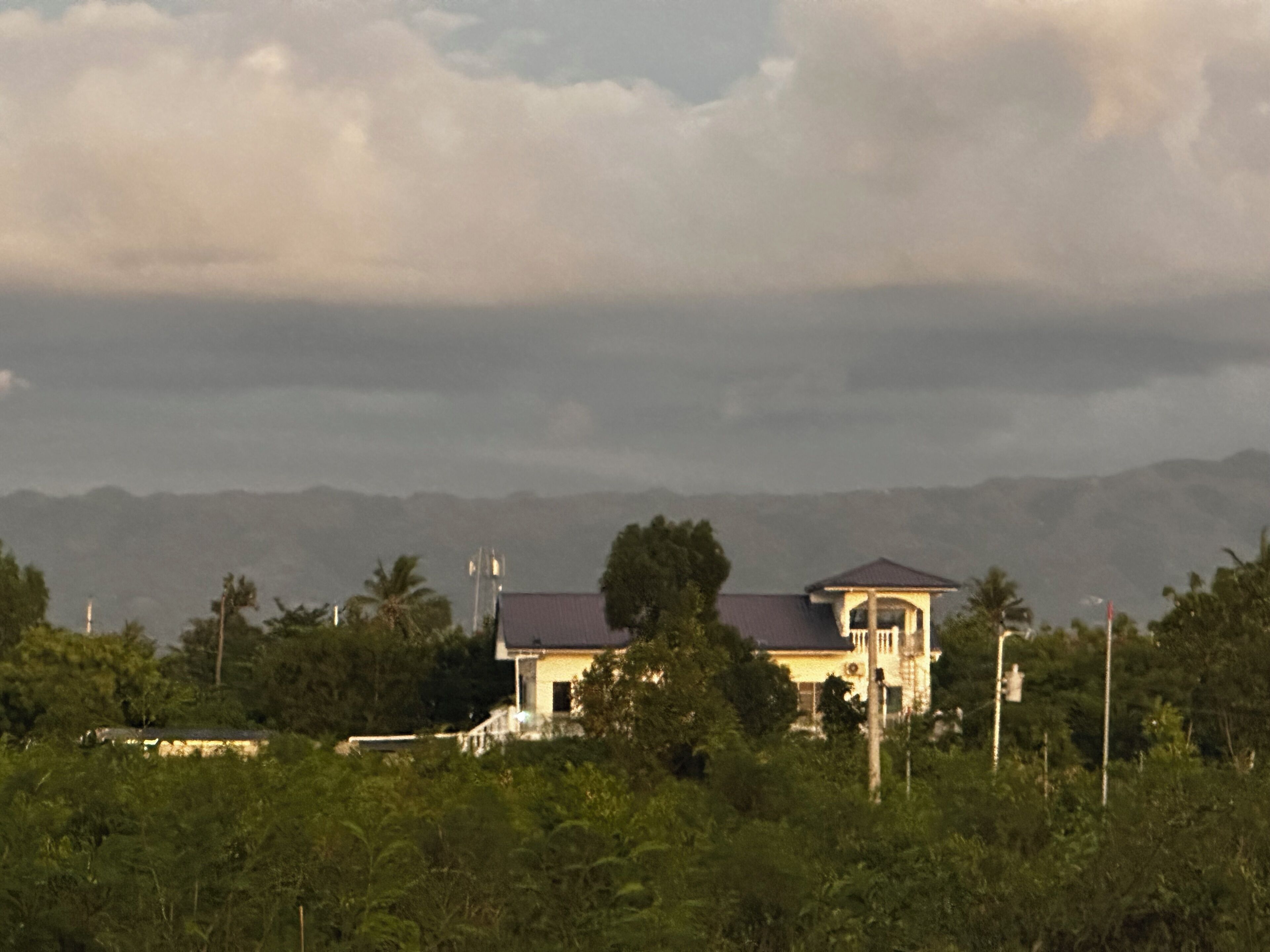 A two-story house with a flat roof, surrounded by dense greenery and a mountainous backdrop.