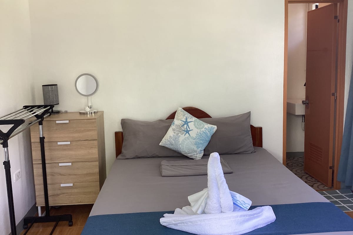 Hotellihuone, jossa on sänky, puinen lipasto, peili ja pyyhe sängyllä.
