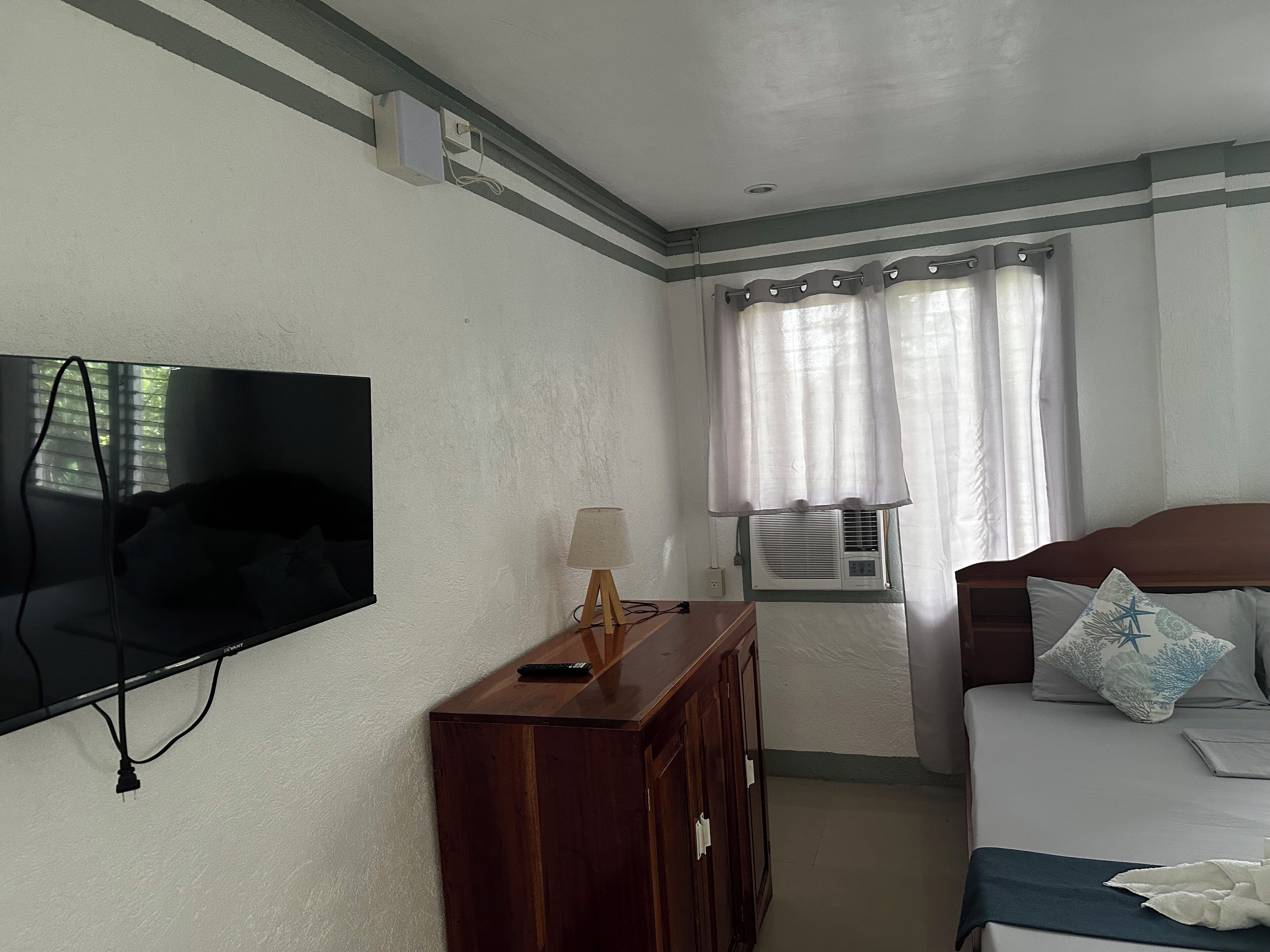 Une chambre d’hôtel équipée d’un téléviseur à écran plat, d’une commode en bois, d’une table de chevet avec une lampe, d’une fenêtre avec des rideaux, d’un climatiseur et d’un lit avec des oreillers.