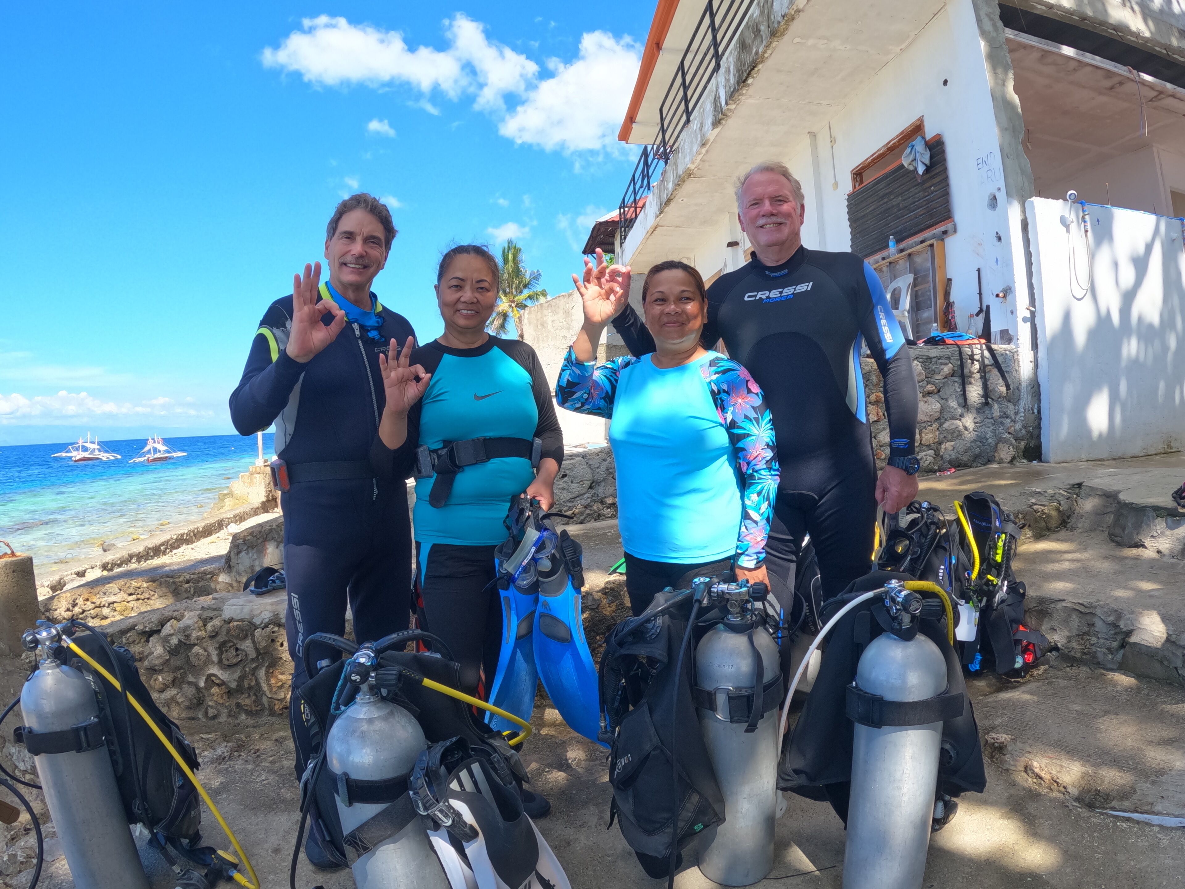 Four people in scuba diving gear standing in front of a building with a view of the ocean and boats in the background.