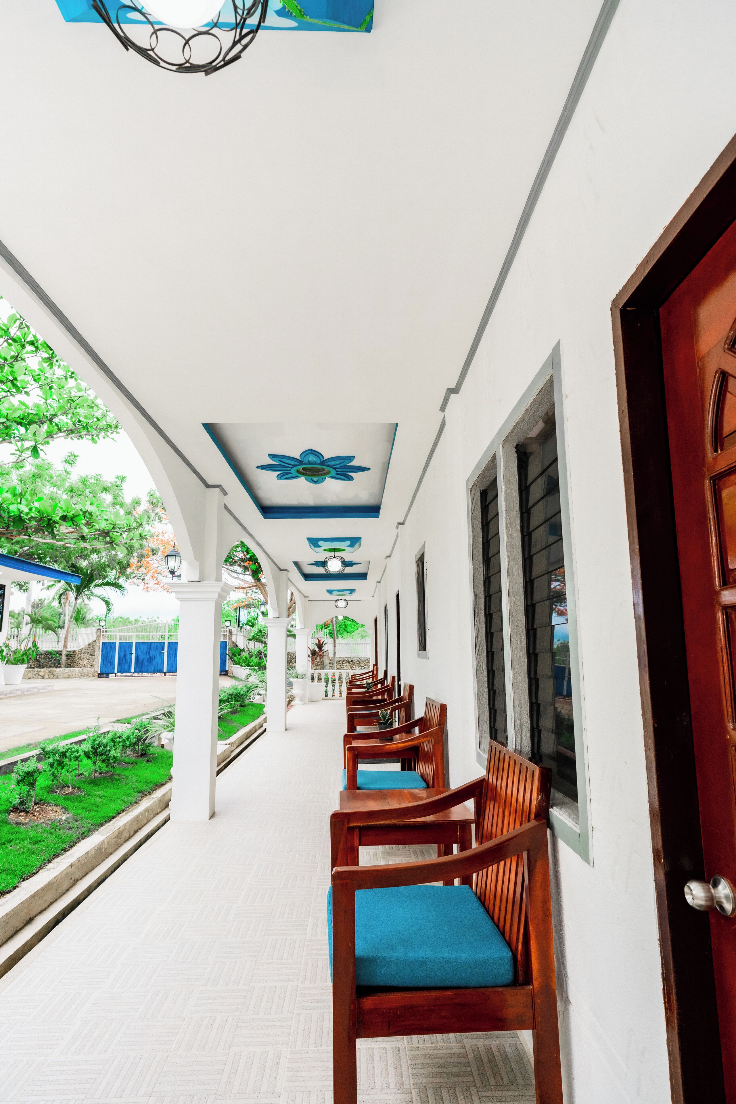 A covered corridor with wooden chairs and tables, white walls, and a view of greenery outside.